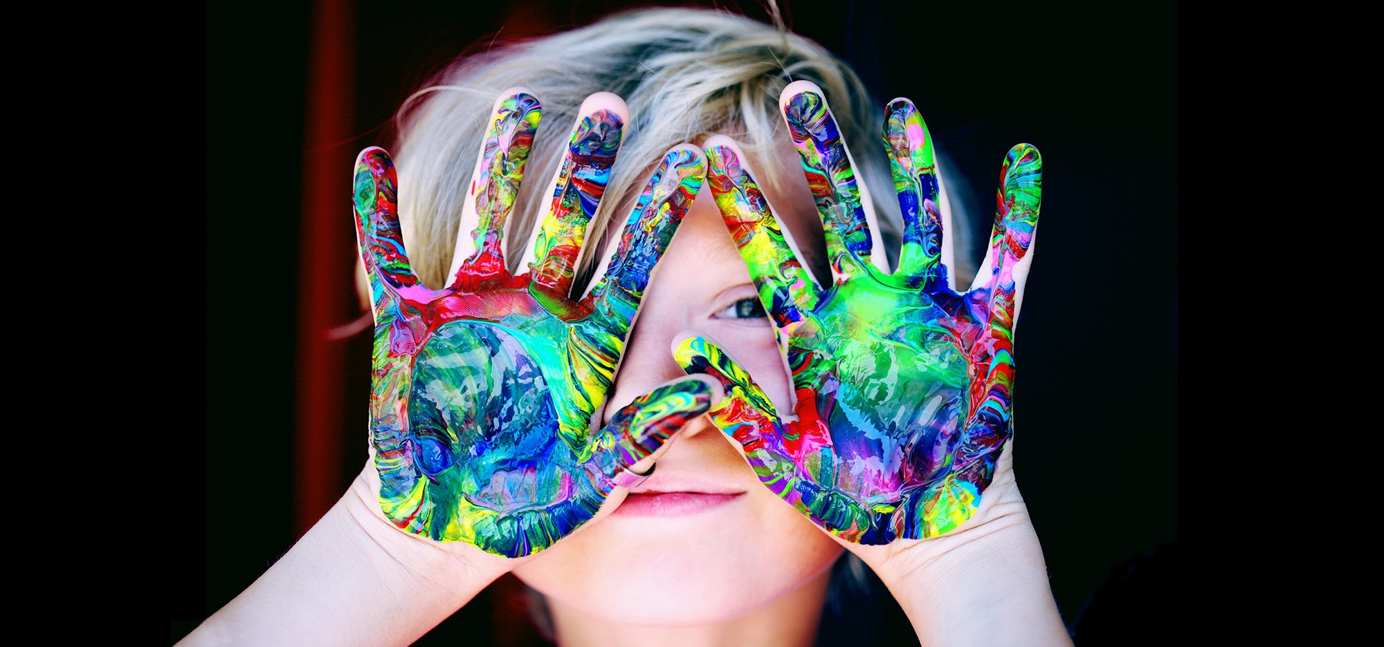 Young child holding their hands covered in paint in front of their face